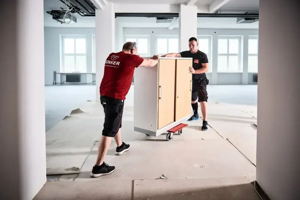 Zwei Mitarbeiter der Umzugsfirma Junker transportieren einen Schrank auf einem Möbelroller durch helle, unfertige Büroräume.