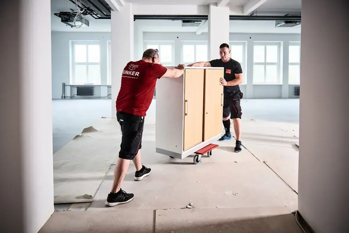 Zwei Mitarbeiter der Junker Umzugsfirma transportieren einen Schrank auf einem Rollbrett durch einen Innenbereich.