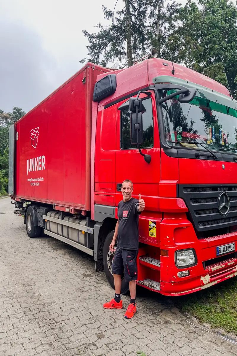 Mitarbeiter der Umzugsfirma Junker steht vor einem roten Junker-Lkw und zeigt Daumen hoch für einen erfolgreichen Bürgergeldumzugs.